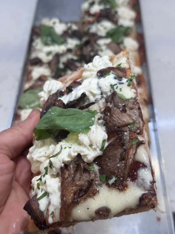 A slice of gourmet pizza from Crustinos Pizza, topped with fresh mushrooms, creamy ricotta, and basil, showcasing the pizzeria's dedication to using high-quality ingredients and traditional New York-style recipes.
